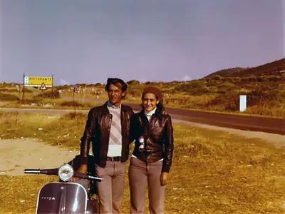 Paola Chiaramonte e Andrea Recchia accanto a una Vespa, su una strada di campagna con un cartello stradale