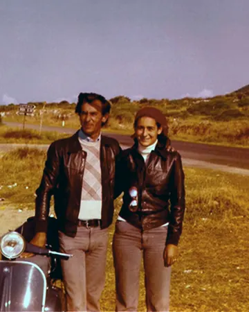 Paola Chiaramonte e Andrea Recchia da giovani, in giacca di pelle, accanto a una Vespa su una strada di campagna